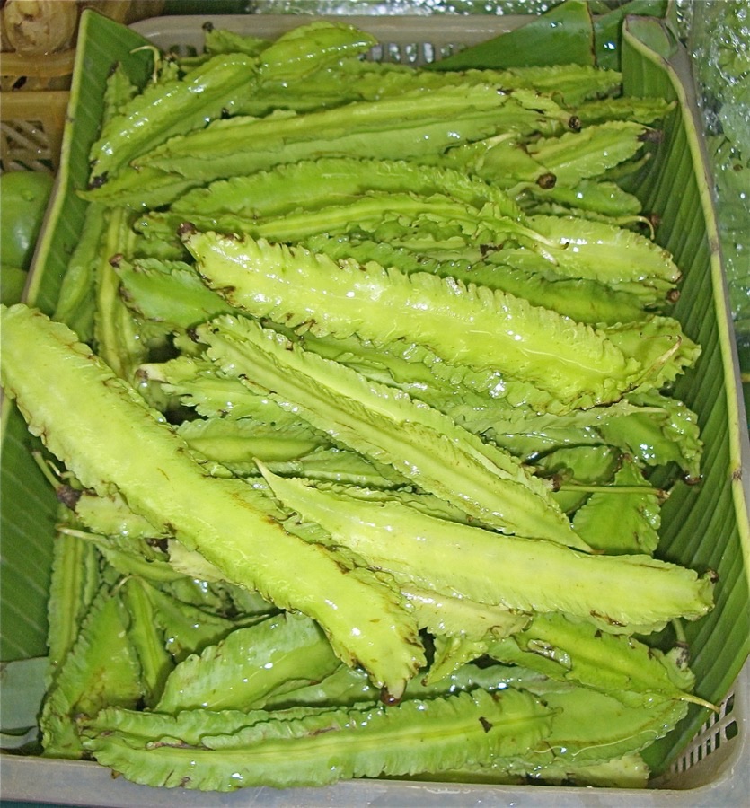 A market crate lined with banana leaf holding many winged bean pods, each four-sided with a frilled green wing running along each corner.